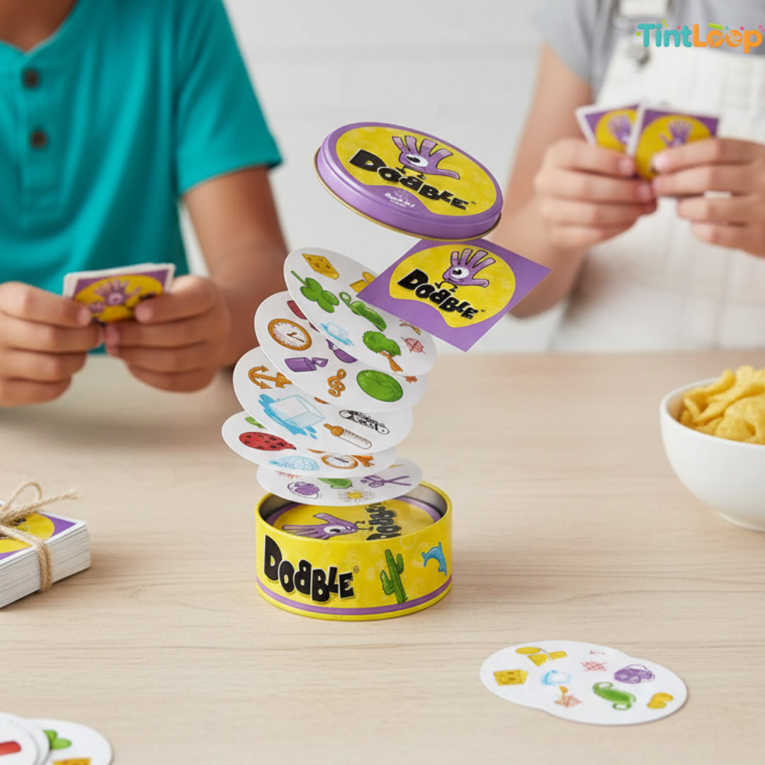 A children's card game called Dooble is shown on a light wood table, with two children holding cards in the background. The game features a yellow cylindrical tin base with a purple lid, and white cards printed with colorful cartoon icons.